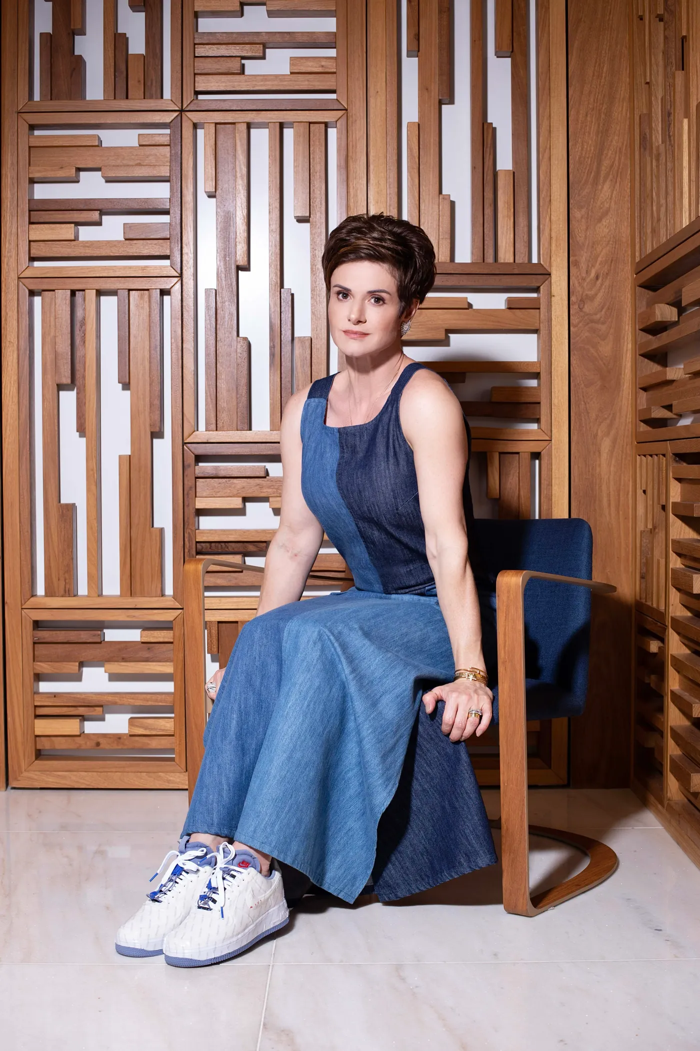 Ká Borges sentada em uma poltrona azul, usando vestido jeans e tênis branco